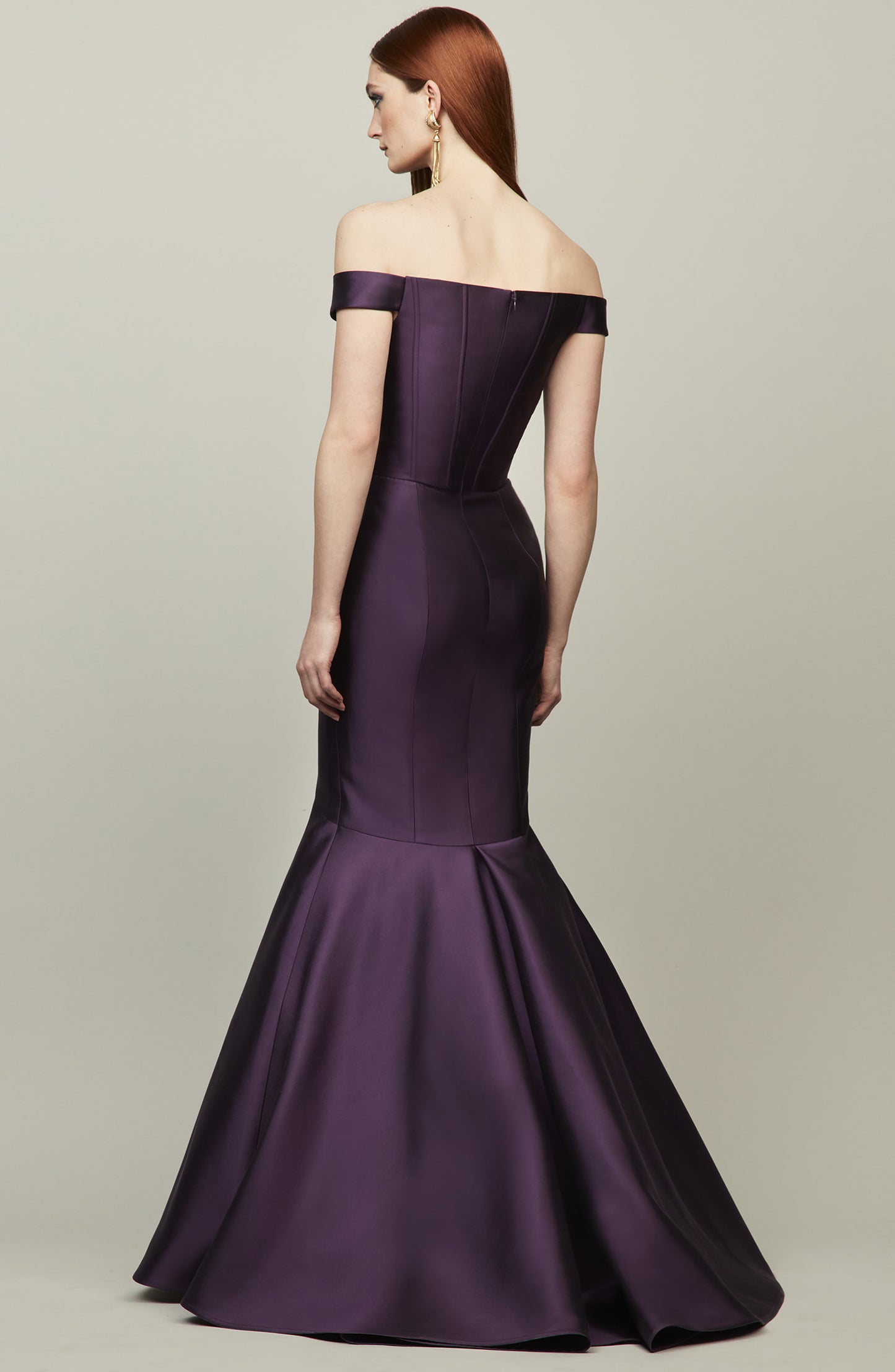 A woman modeling an off-the-shoulder mermaid gown in purple shown from the back against a neutral light gray studio background.