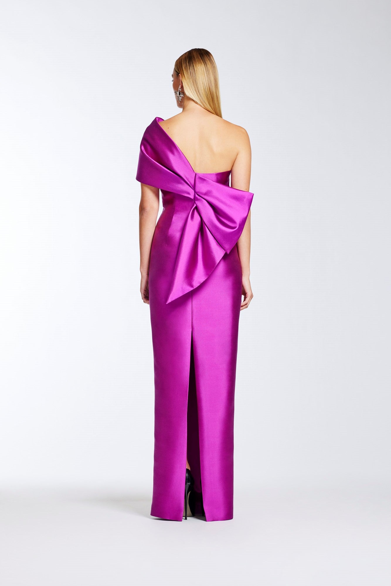 A woman stands with her back to the camera wearing a fuchsia one-shoulder satin gown with a large bow detail at the back, photographed against a light gray studio background.