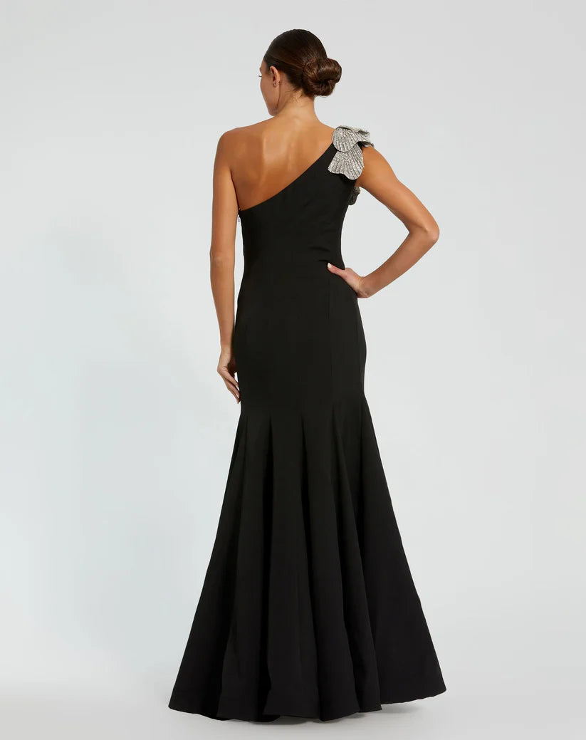 Model wearing Mac Duggal 11810 one-shoulder trumpet gown with flower detail. Back View.