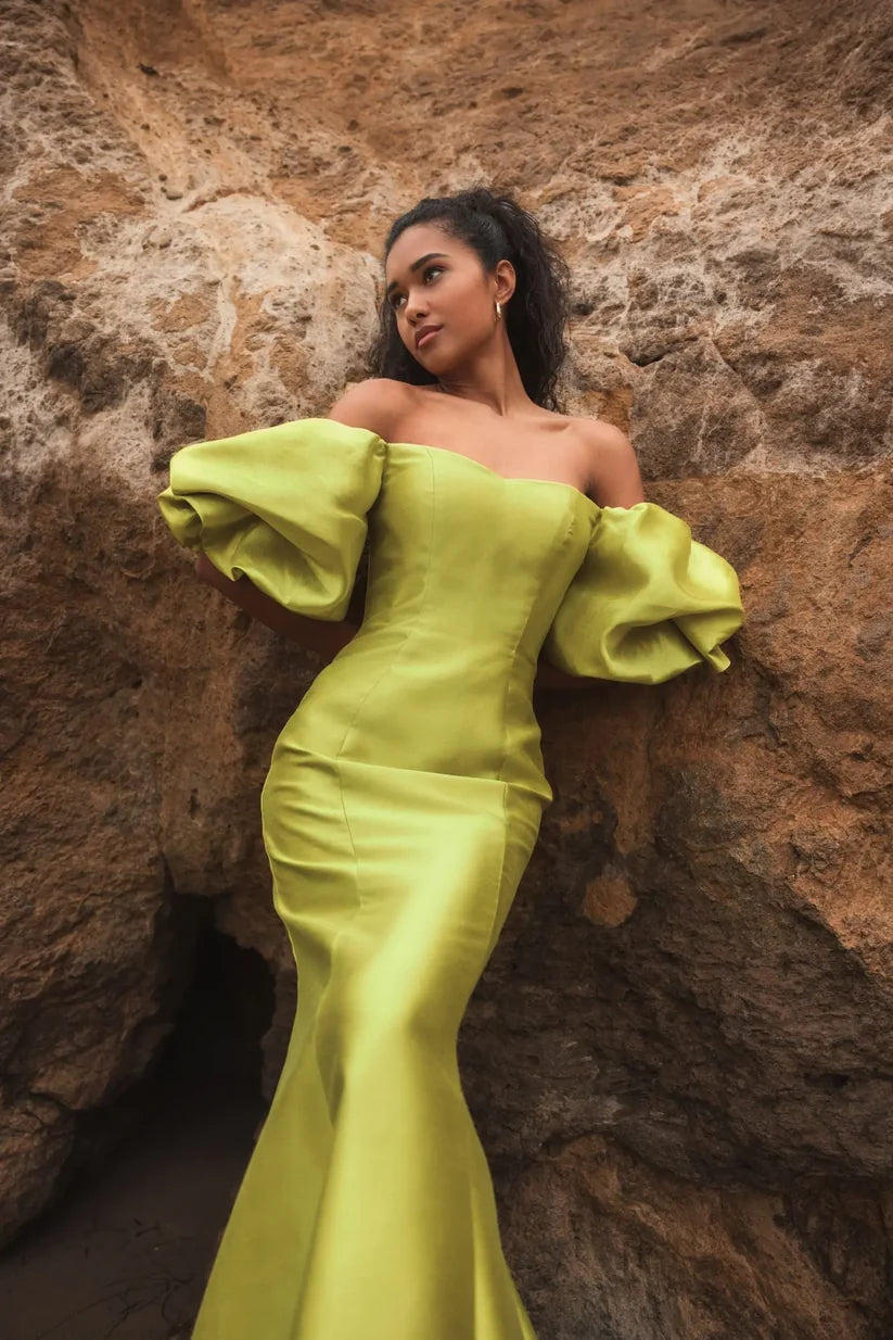Model wearing Mac Duggal 50677 off-the-shoulder satin gown with puff sleeves, mermaid silhouette, and sweeping train. Model is outside wearing the dress in the color chartreuse.