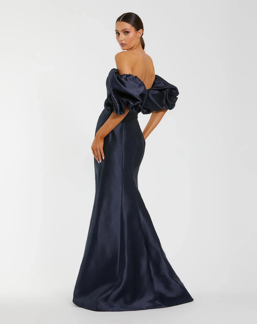 Model wearing Mac Duggal 50677 off-the-shoulder satin gown with puff sleeves, mermaid silhouette, and sweeping train. Model is wearing the color midnight. Back 3/4 view.