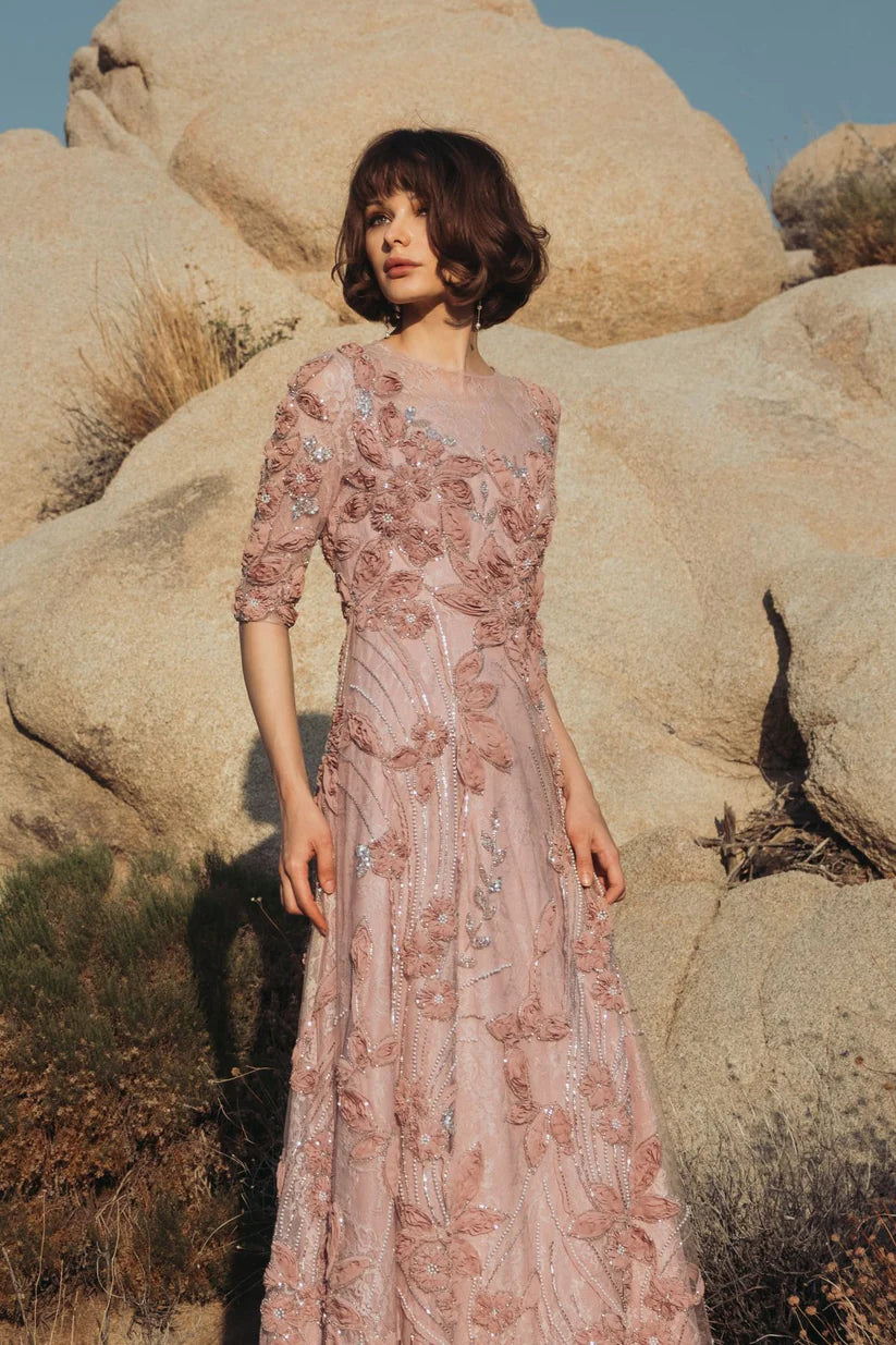 Model wearing Mac Duggal 69016 gown with illusion neckline, 3/4 sheer sleeves, 3D rosettes, and a flowing embellished skirt. Model outside wearing the dress i the color mocha.