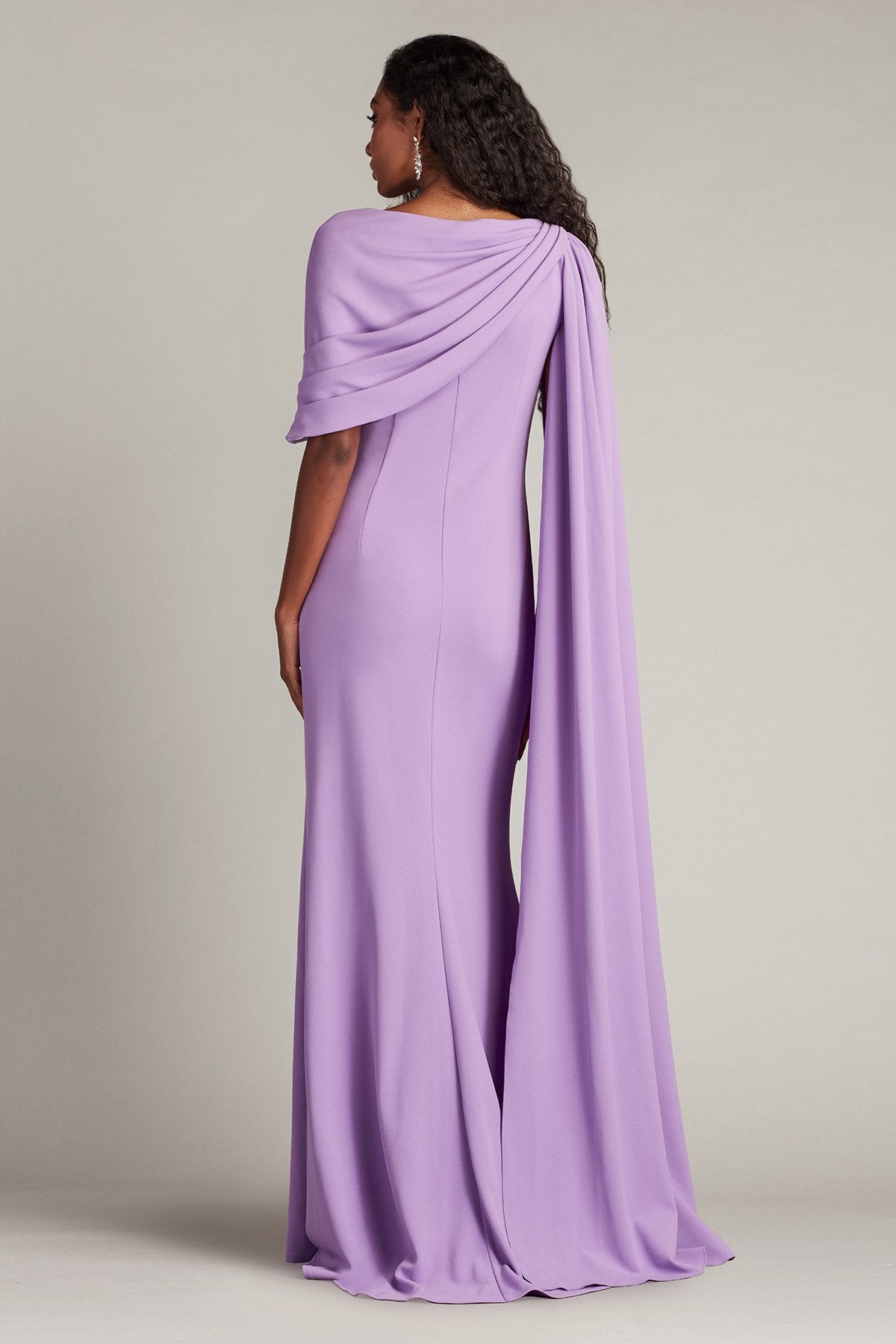 Model wearing Tadashi Shoji BOS25345L gown with one-shoulder cape, pleated bodice, and side slit, shown in front and back views.