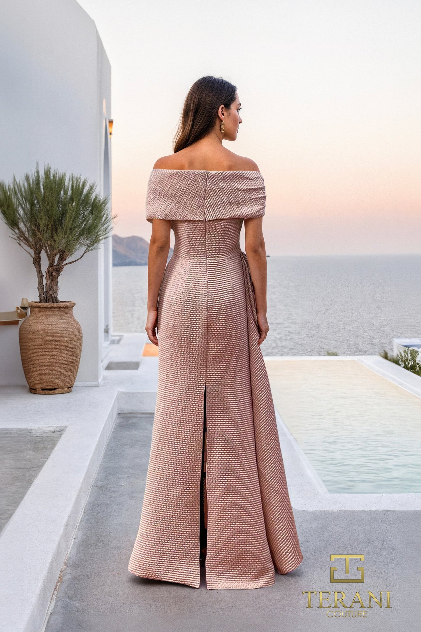 Model wearing Terani Couture 261M6679 off-shoulder sheath gown with side gathered detail and embellishment, posing outdoors by the sea.