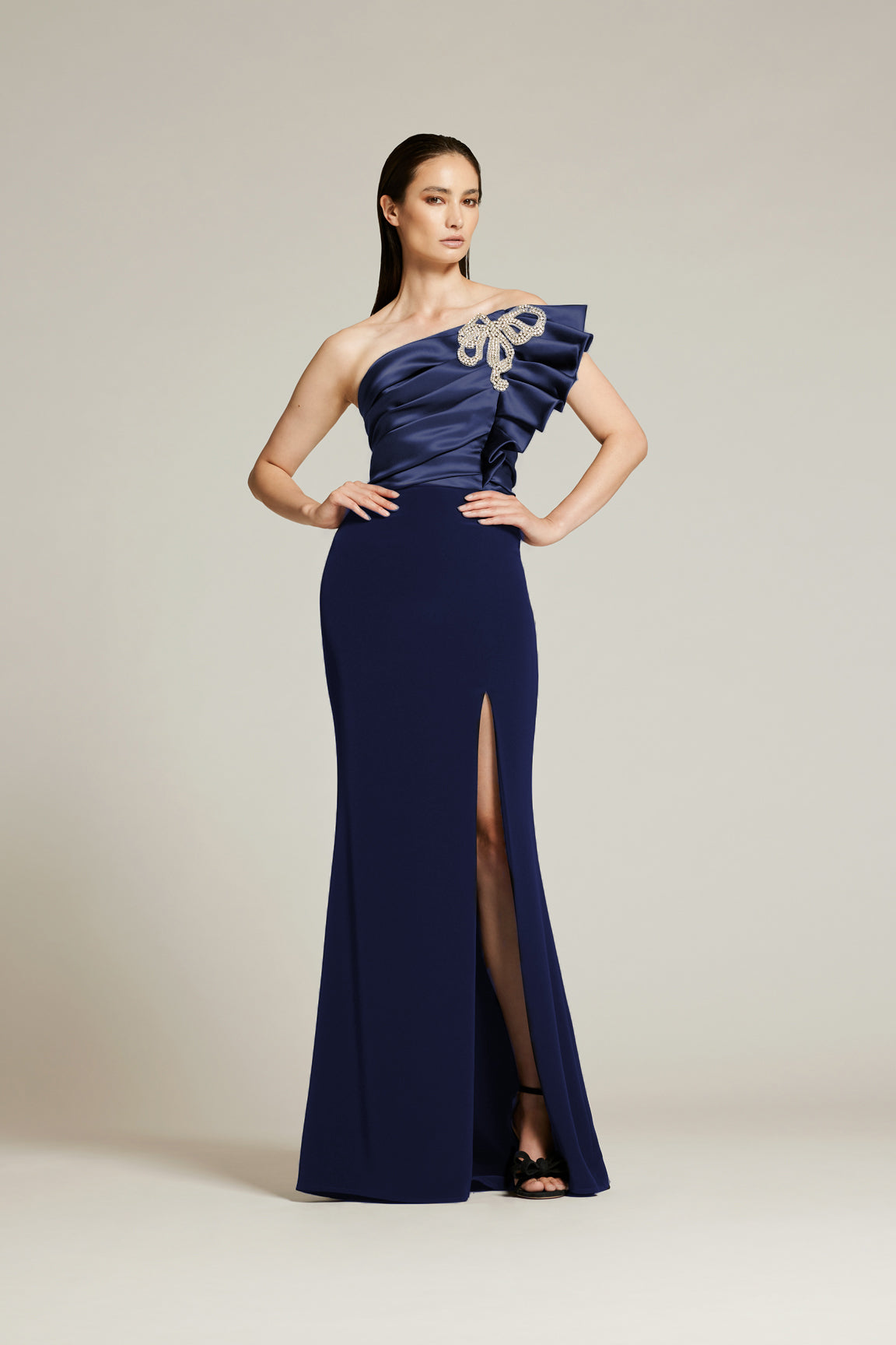 A woman modeling a navy blue, strapless, draped bodice gown with a crystal bow applique, featuring a fit and flare silhouette with front side slit, and a hidden back zipper.