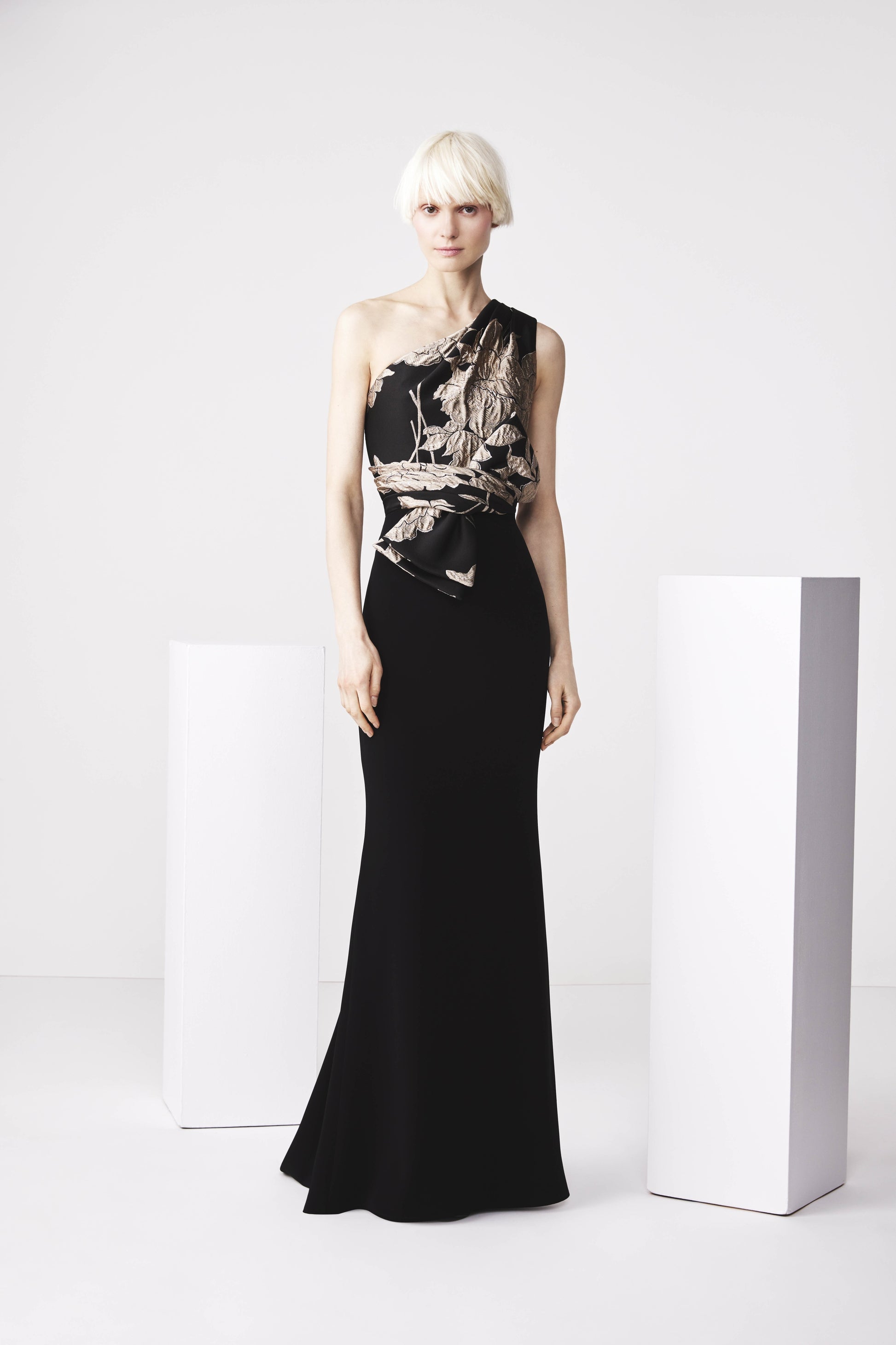 A model wearing a black, one-shoulder gown with a contrast draped bodice and a bow at the waist, standing next to white column-like props.