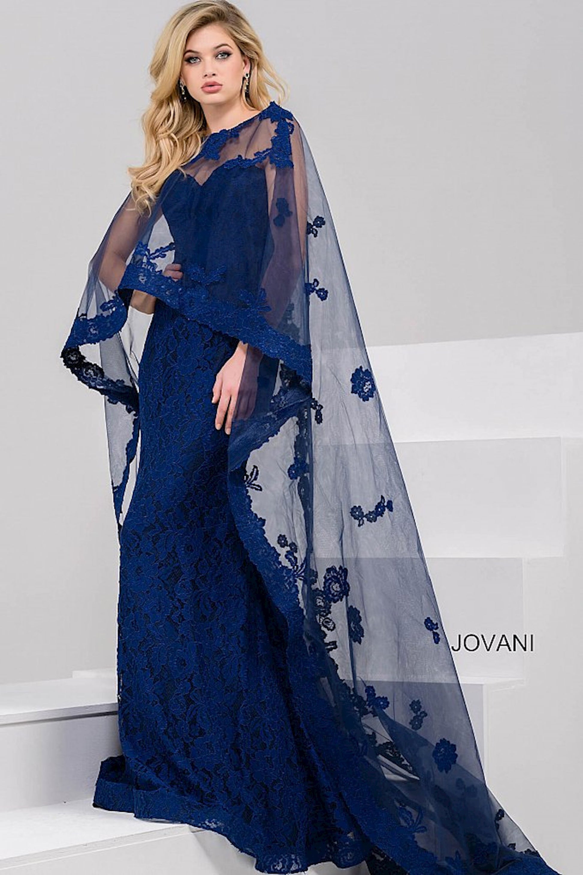 Jovani 39769 Evening Gown with Lace and Sheer Cape Overlay – A floor-length lace gown featuring a high neckline and dramatic sheer cape adorned with floral appliqués.
