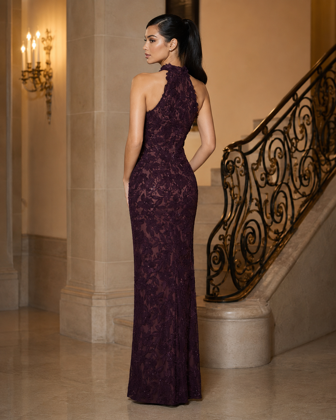 A woman stands in a grand interior hallway wearing a sleeveless, halter-neck floor-length gown made of aubergine color lace. The dress has a fitted silhouette and is shown from the back, with a staircase and stone columns in the background.