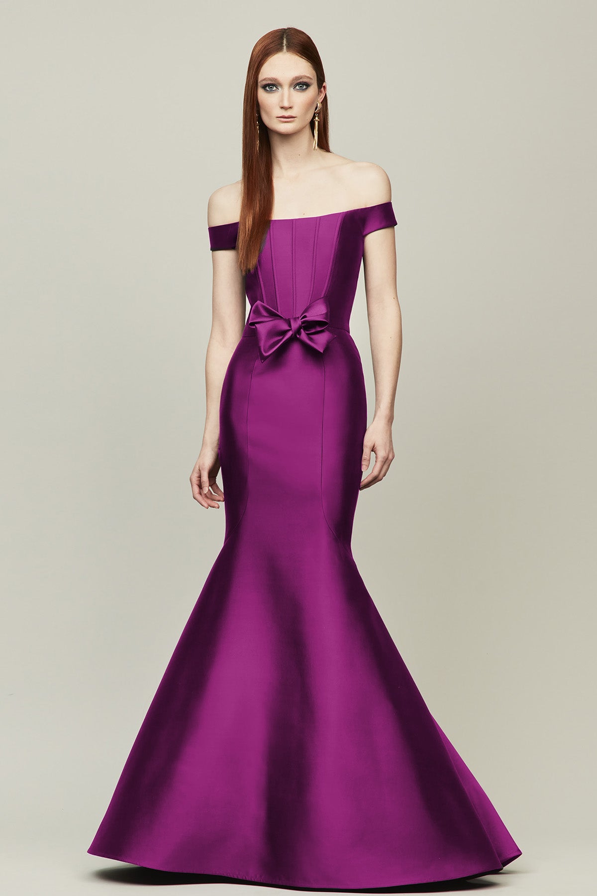 A woman modeling a floor-length off-the-shoulder mermaid gown in grape color featuring a large bow at the waist, set against a solid light gray studio background.