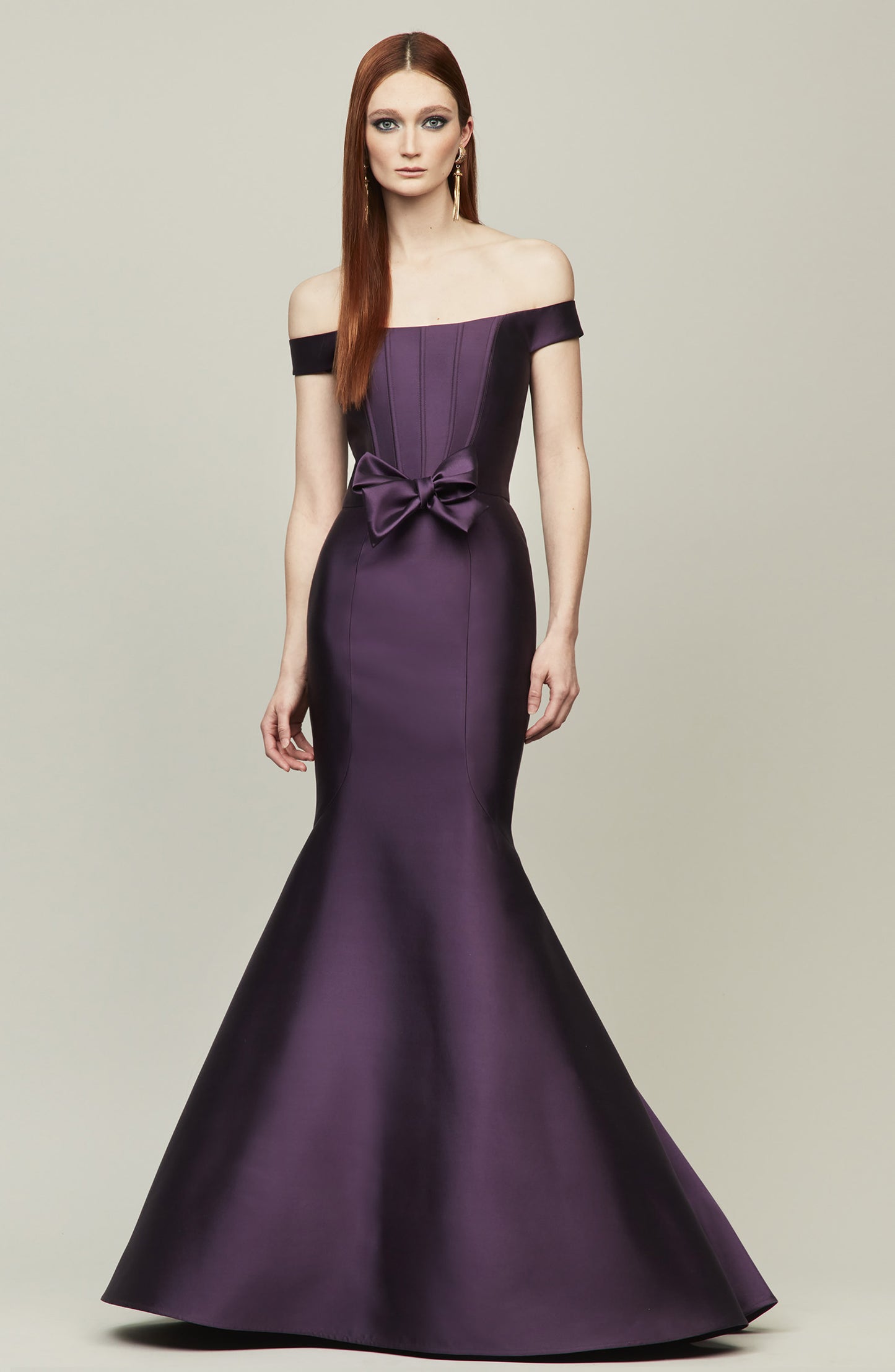A woman modeling an off-the-shoulder floor-length gown in purple featuring a structured bodice, a large bow at the waist, and a flared mermaid skirt, against a solid light gray studio background.