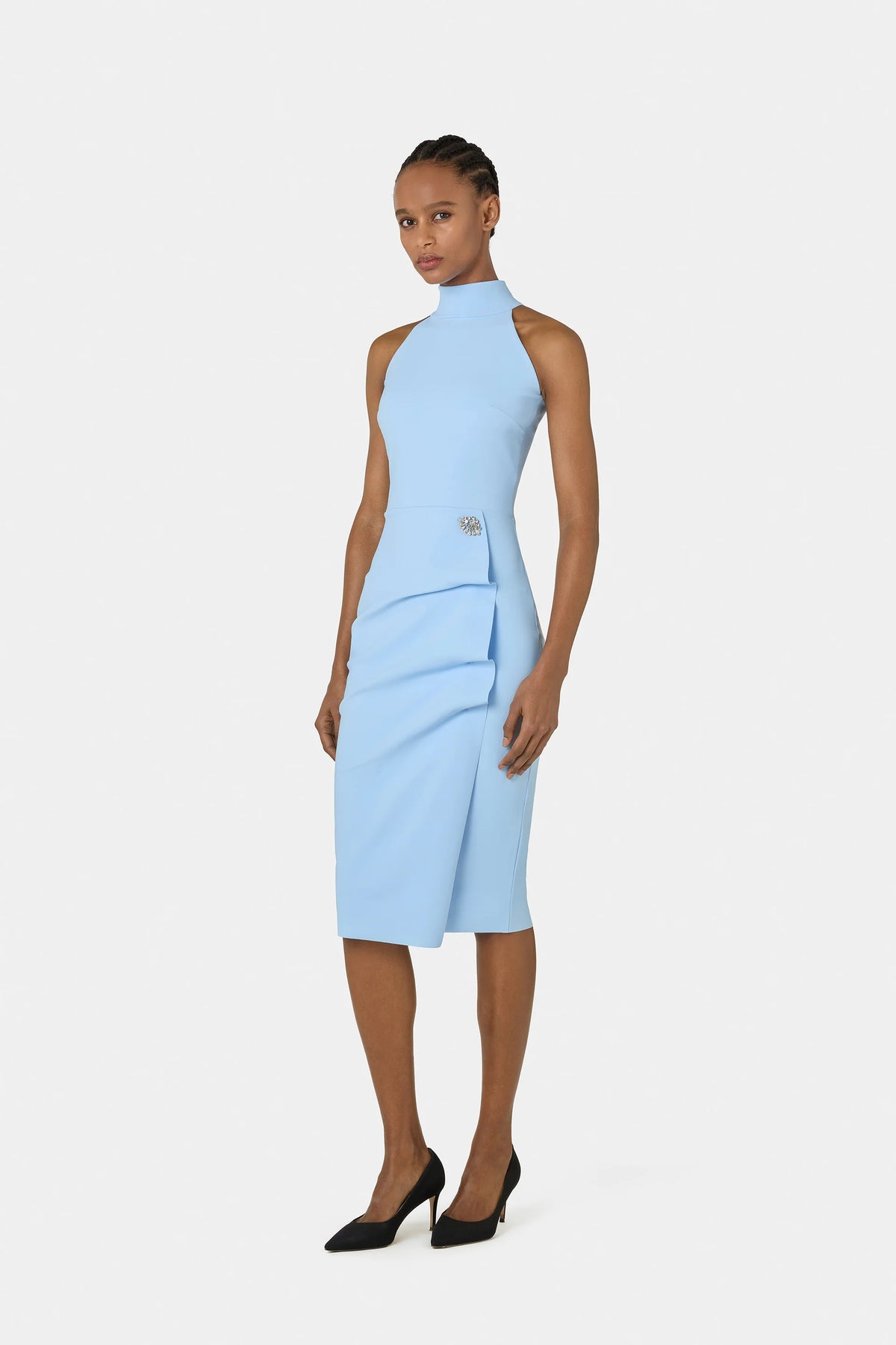 A model wearing a light blue sleeveless mock-neck midi dress with a wrap-style skirt and a small gold button detail at the waist, paired with black pointed-toe heels, against a light gray studio background.