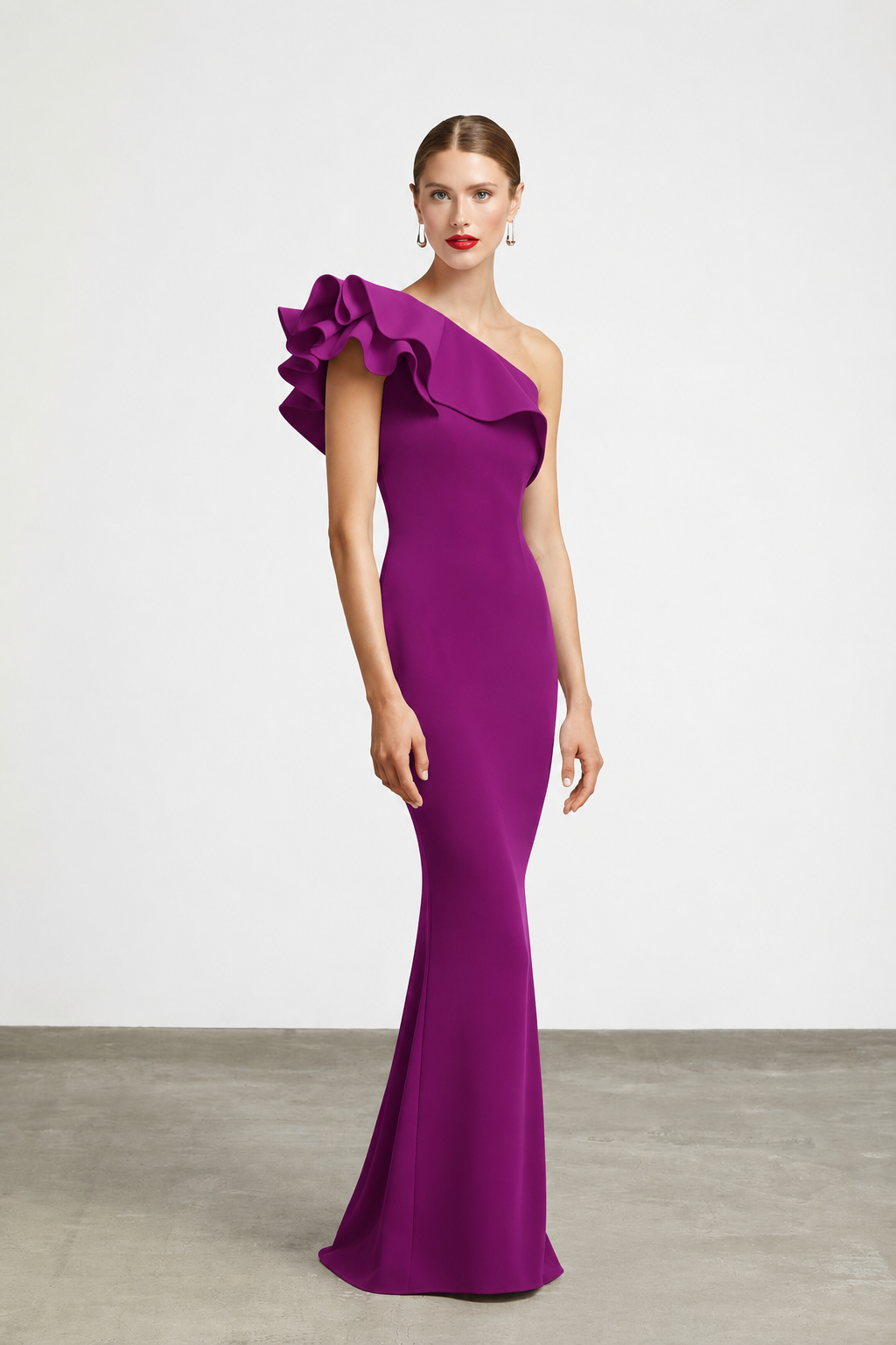 A woman modeling a floor-length, one-shoulder magenta gown with a dramatic ruffled shoulder detail, photographed in a studio with a light gray wall and concrete floor.
