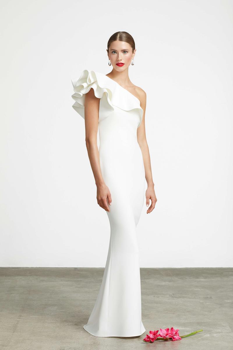 A woman models a floor-length white one-shoulder gown with a dramatic ruffled shoulder detail, standing against a plain light gray studio background with a concrete floor and a small pink flower at her side.
