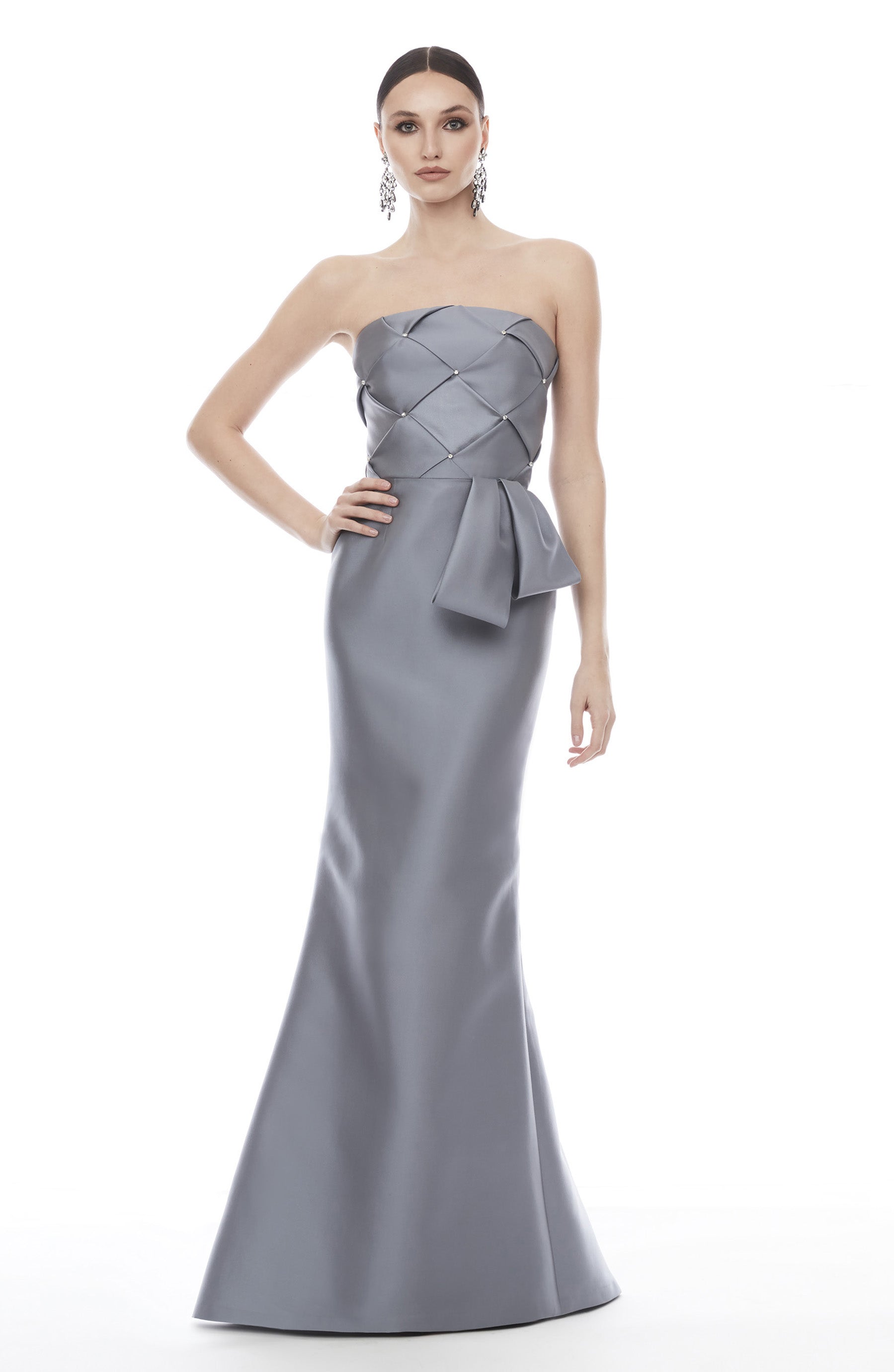 A woman modeling a strapless floor-length gown in a cool light gray or silver satin-like fabric. The dress has a structured, quilted-looking bodice with a large bow detail at the waist and a fitted mermaid silhouette.