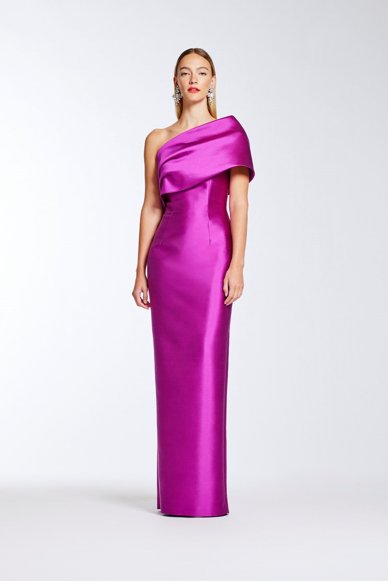 A woman modeling a floor-length, one-shoulder satin gown in vivid fuchsia against a clean light gray studio background.