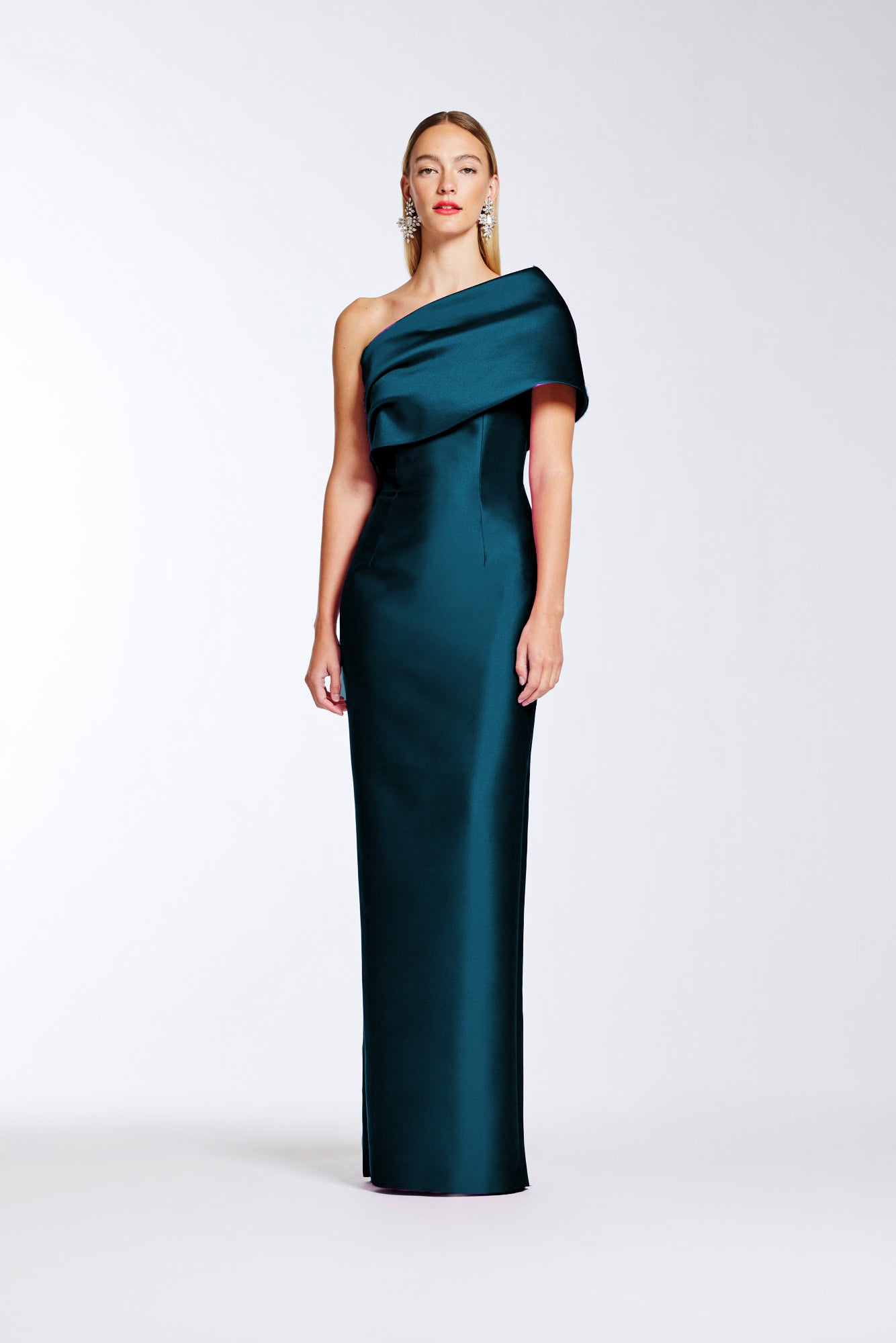 A woman modeling a floor-length, one-shoulder satin gown in deep teal green against a clean light gray studio background.