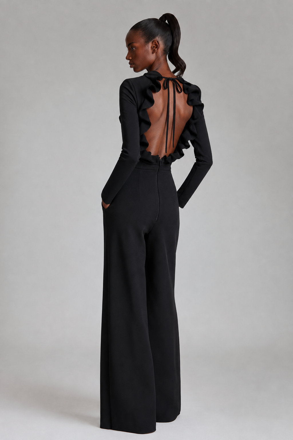 A model shown from the back wearing a black long-sleeve wide-leg jumpsuit with a ruffled open back and tie details, photographed against a neutral light gray studio background.