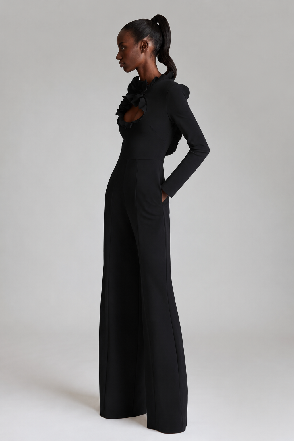 A model in profile wearing a black long-sleeve jumpsuit with a ruffled collar and wide-leg pants, photographed against a neutral light gray studio background.