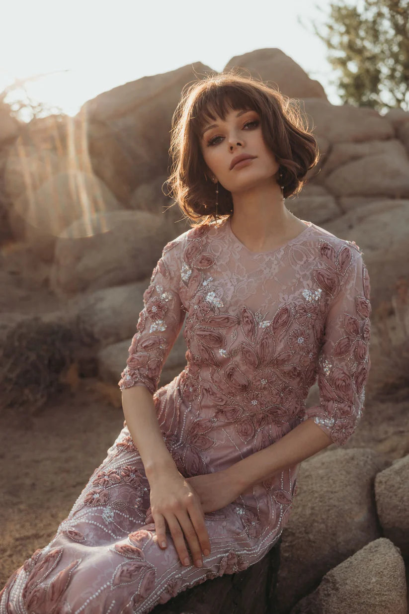 Model wearing Mac Duggal 69016 gown with illusion neckline, 3/4 sheer sleeves, 3D rosettes, and a flowing embellished skirt.  Model outside wearing the dress i the color mocha.