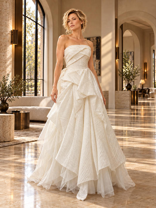 A woman models a strapless white wedding gown with a structured crinkled bodice and a layered, cascading tulle skirt in a bright, elegant interior hallway.