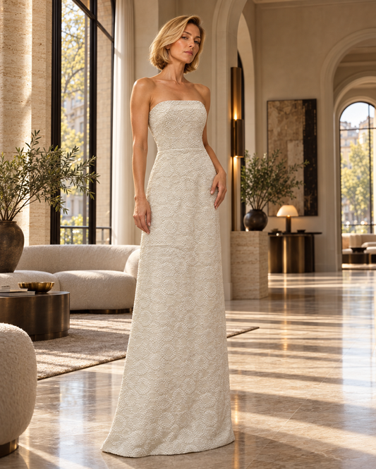 A woman models a strapless, floor-length ivory lace gown in a luxurious, sunlit modern interior with arched windows and beige stone walls.