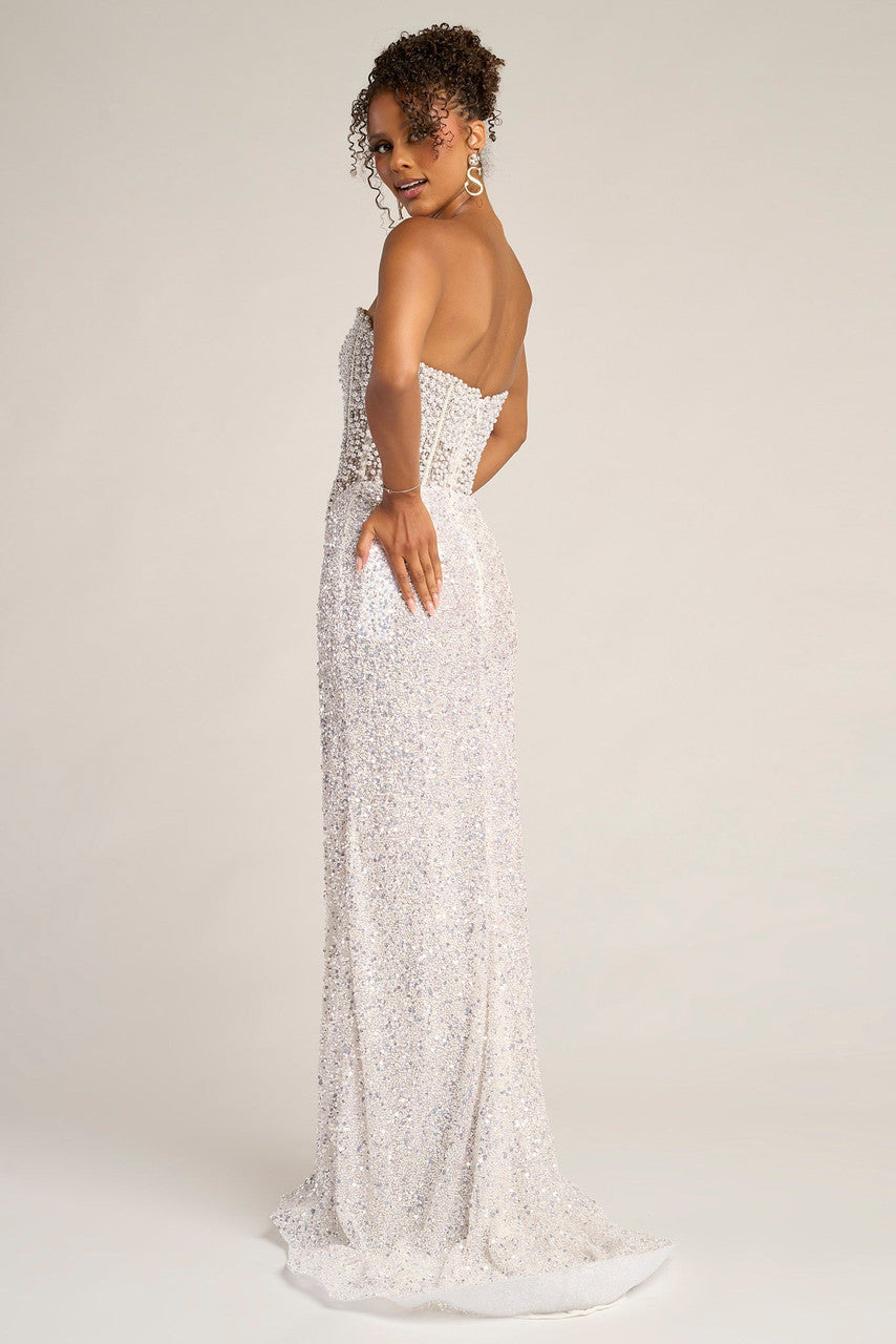 Woman wearing a sparkling white strapless gown with a high slit against a beige curtain backdrop.