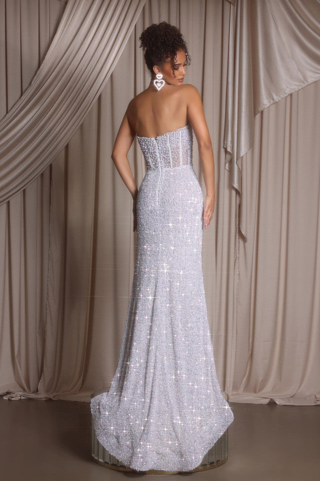 Woman wearing a sparkling white strapless gown with a high slit against a beige curtain backdrop.