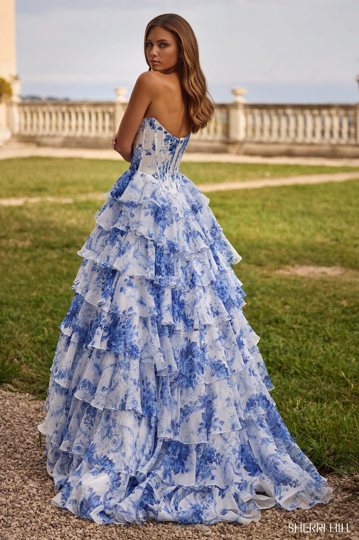 Sherri Hill 57250 Floral Corset Ball Gown – A strapless blue floral print satin gown featuring a structured corset bodice, cascading ruffle skirt, and a high slit for an elegant, romantic look.