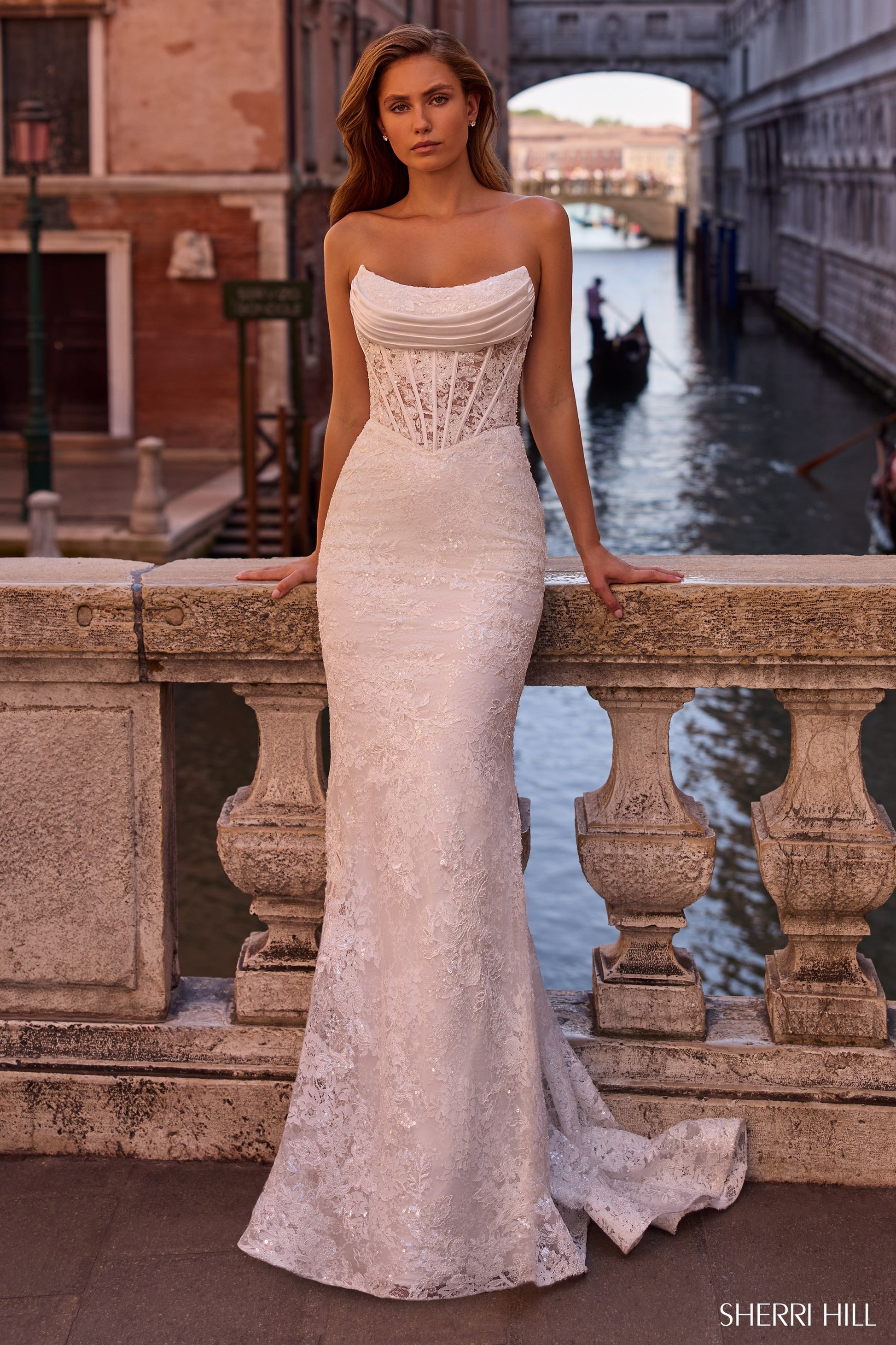 Woman in a strapless lace gown standing by a canal with gondolas in the background
