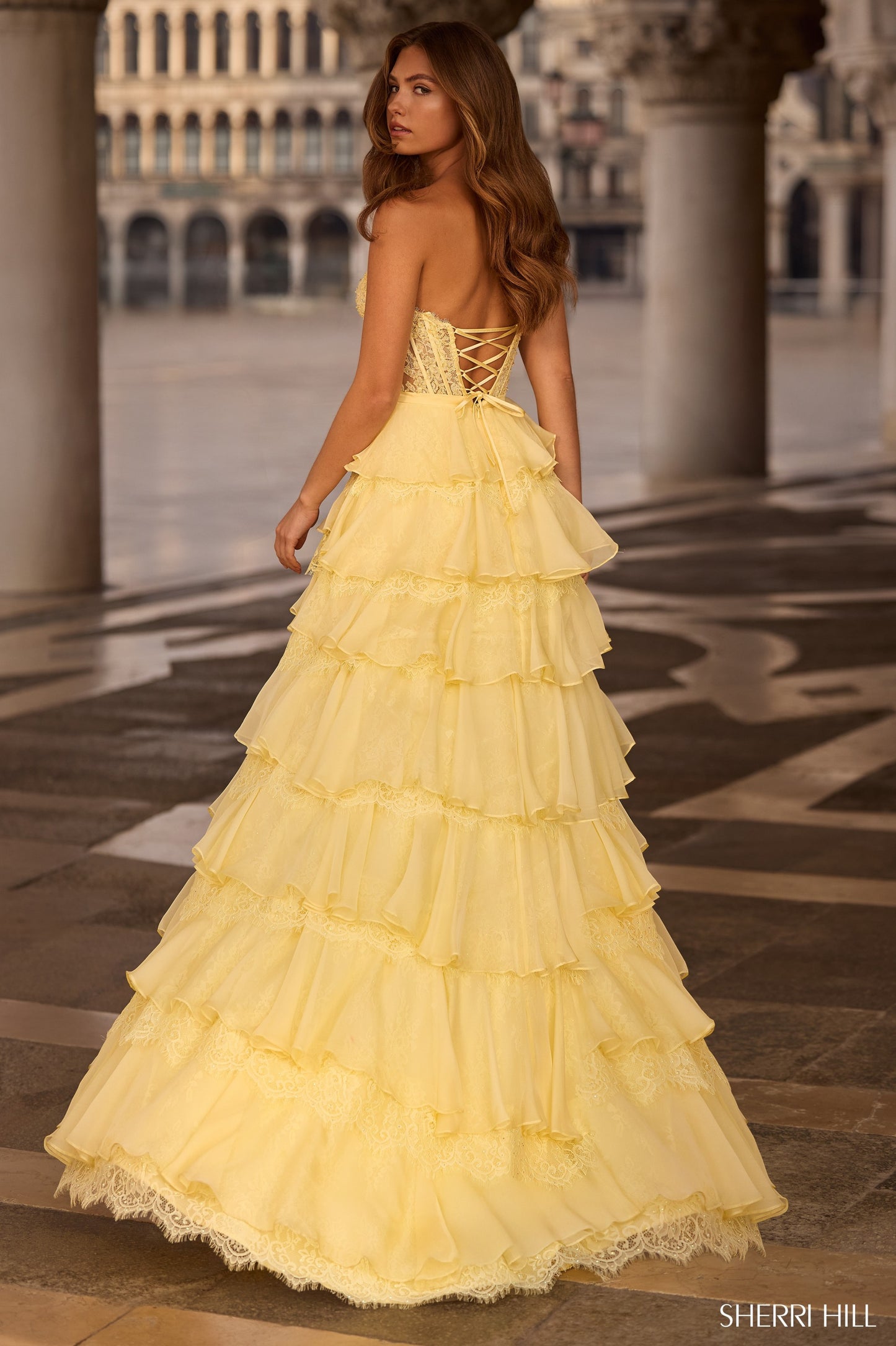 Woman in a yellow evening gown standing on a patterned floor with architectural background