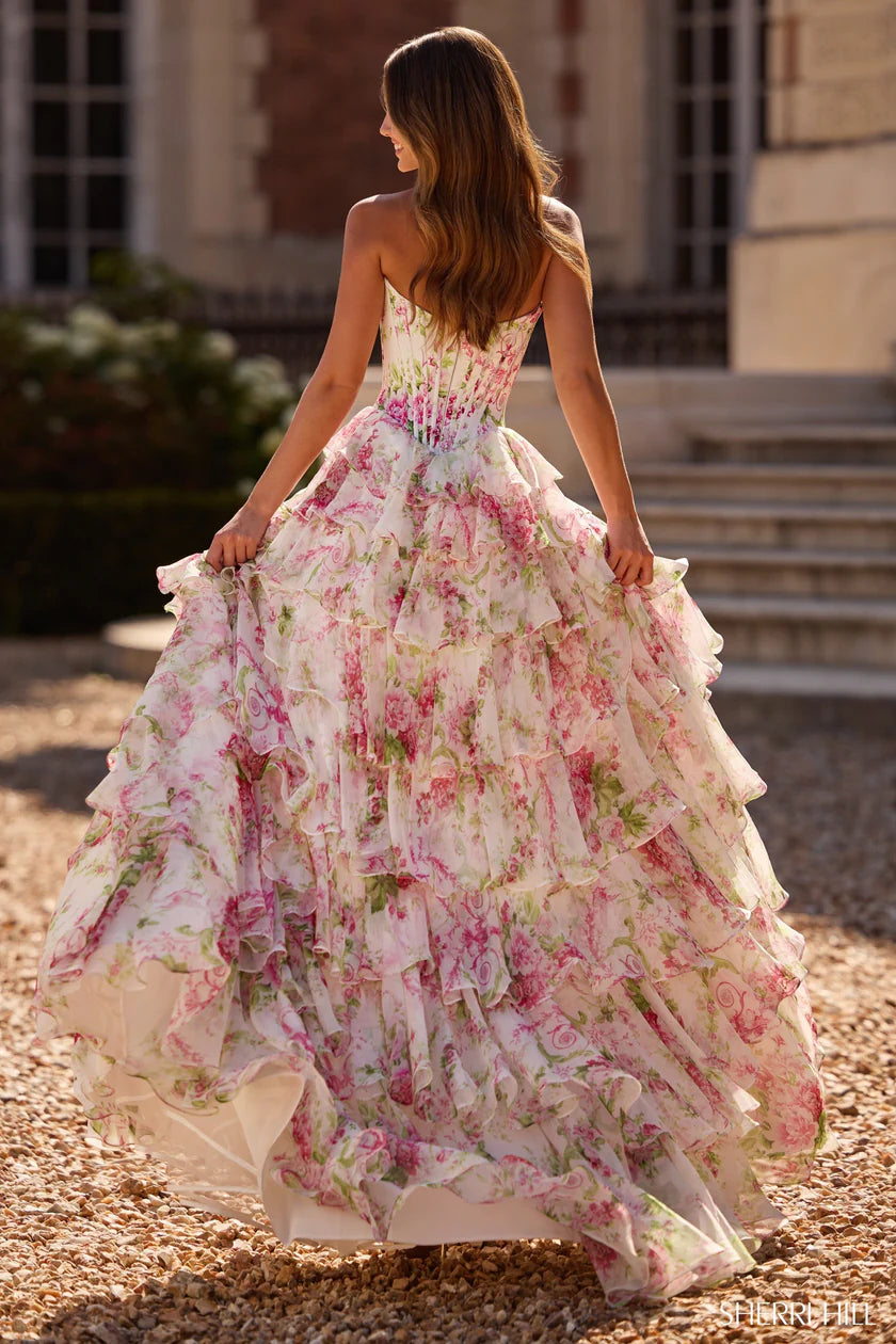 Model wearing Sherri Hill 57693 strapless floral gown with ruffle-layered skirt and slit, posing outdoors in elegant sunlight.