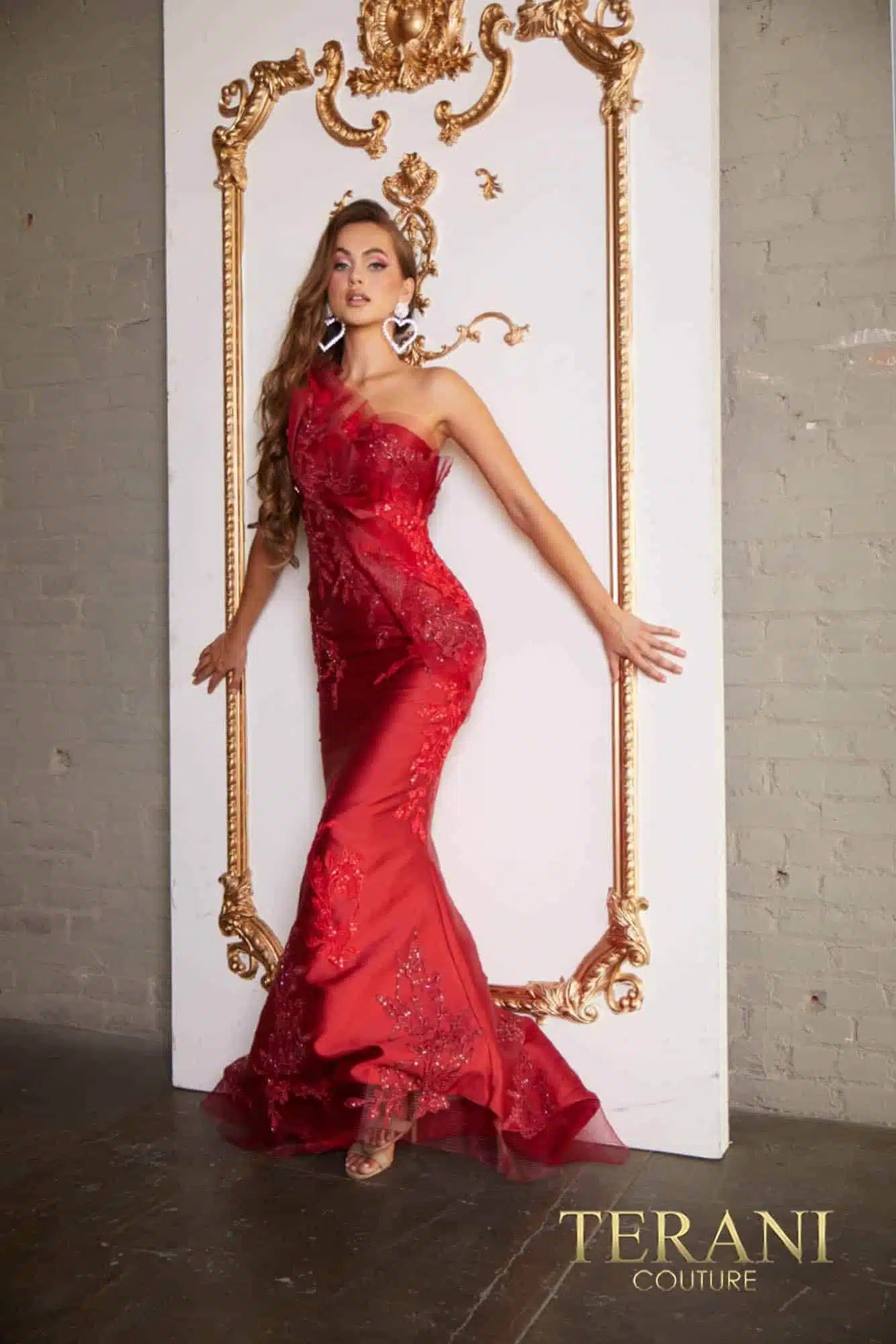 Woman in a ruby color dress standing next to an ornate gold frame with 'Terani Couture' branding.
