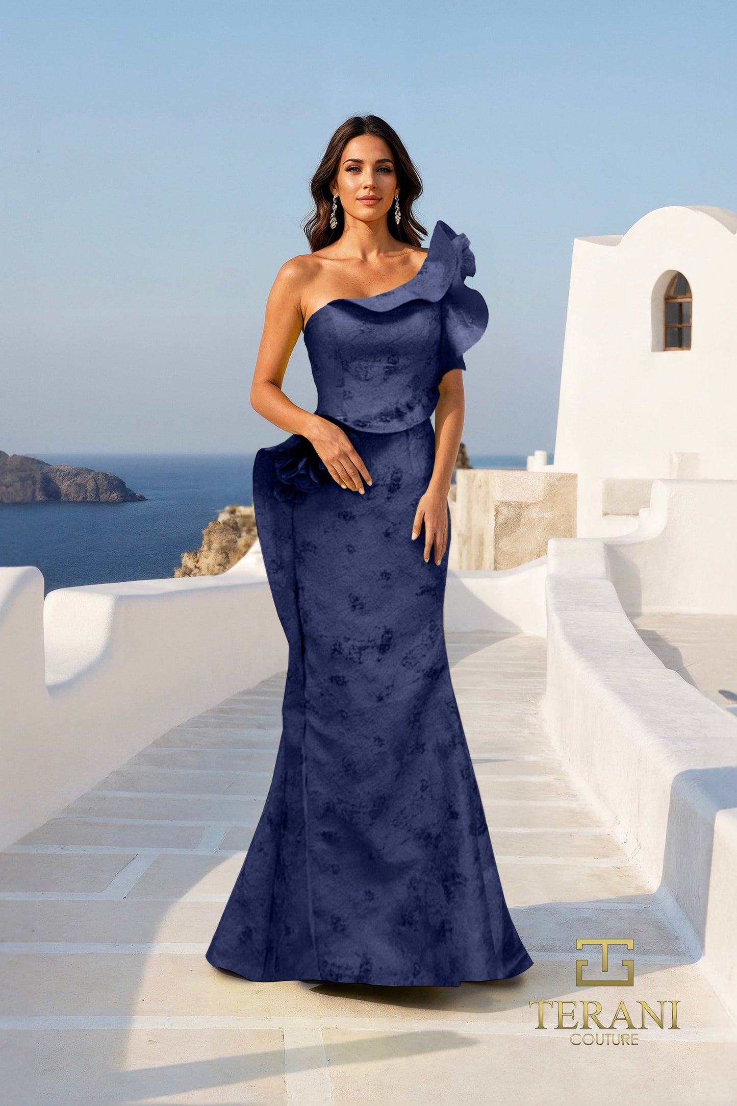 Woman in a blue evening gown standing on a white staircase with ocean view, Terani brand visible